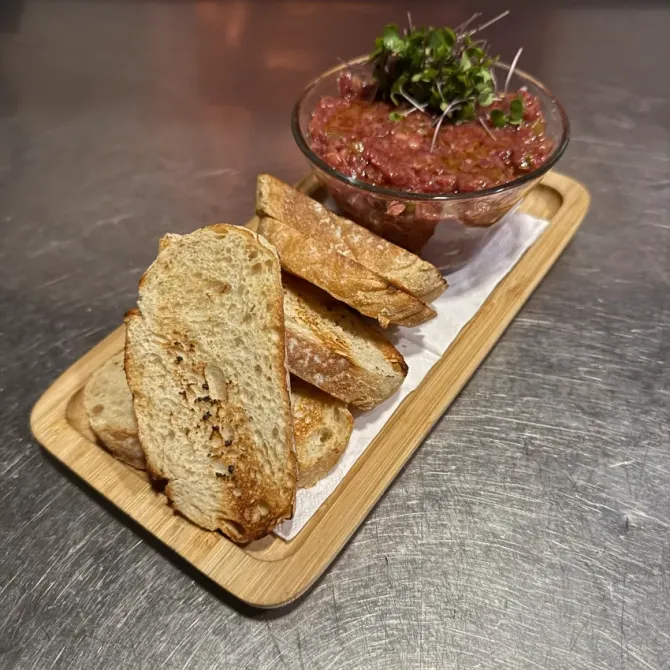 Rebanadas de pan rústico y un tazón de salsa roja adornado con microvegetales en una bandeja de madera.