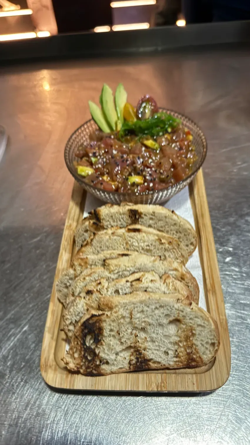 Bandeja de madera con rebanadas de pan y un tazón de salsa marrón con guarnición verde sobre una mesa.