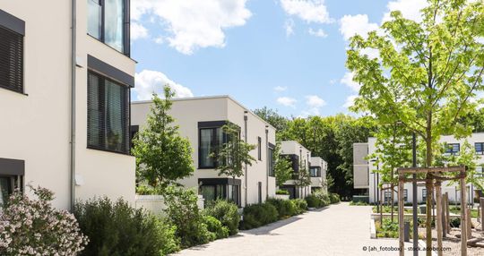 Moderne Wohnanlage mit weißen, kubischen Häusern, grünen Gärten und einem gepflasterten Weg unter blauem Himmel.