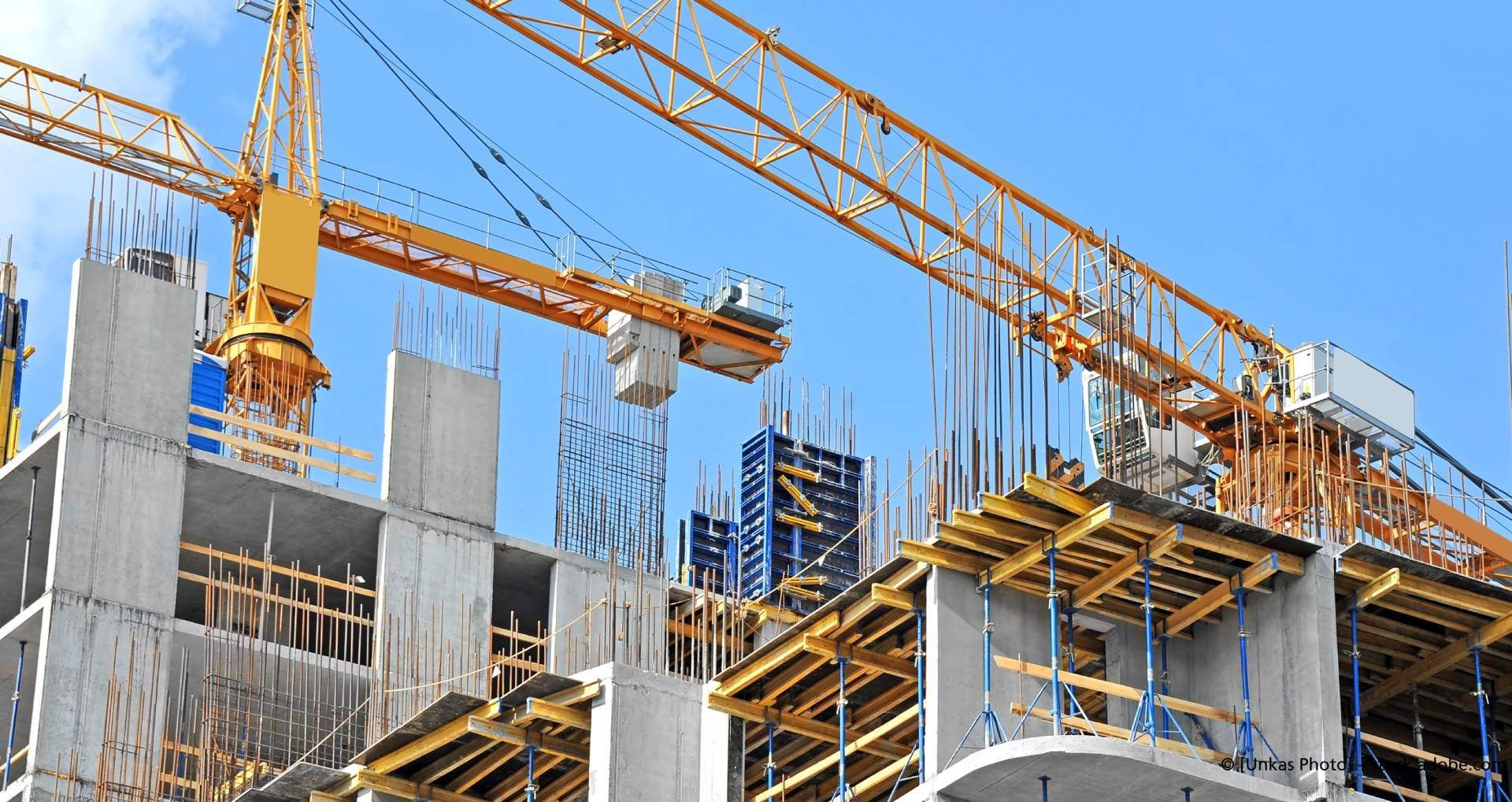 Eine aktive Großbaustelle für ein mehrstöckiges Gebäude aus Beton mit zwei großen gelben Turmdrehkranen vor einem klaren blauen Himmel.