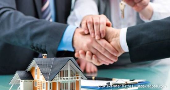 Nahaufnahme eines Modellhauses, im Hintergrund schütteln zwei Leute sich die Hände und ein Schlüssel wird gehalten.