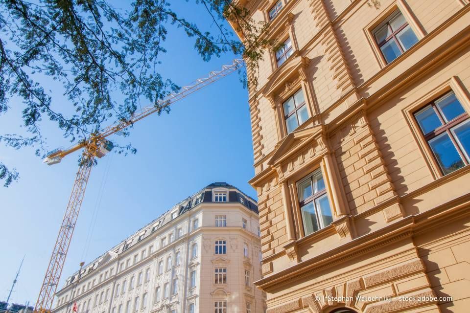 Gebäude mit reich verzierten Fassaden, ein Kran vor blauem Himmel, Baumzweige oben links.