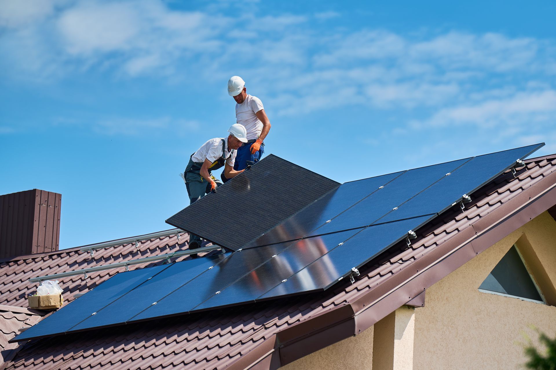 Zwei Arbeiter mit Schutzhelmen installieren Solarpaneele auf einem Dach vor blauem Himmel.