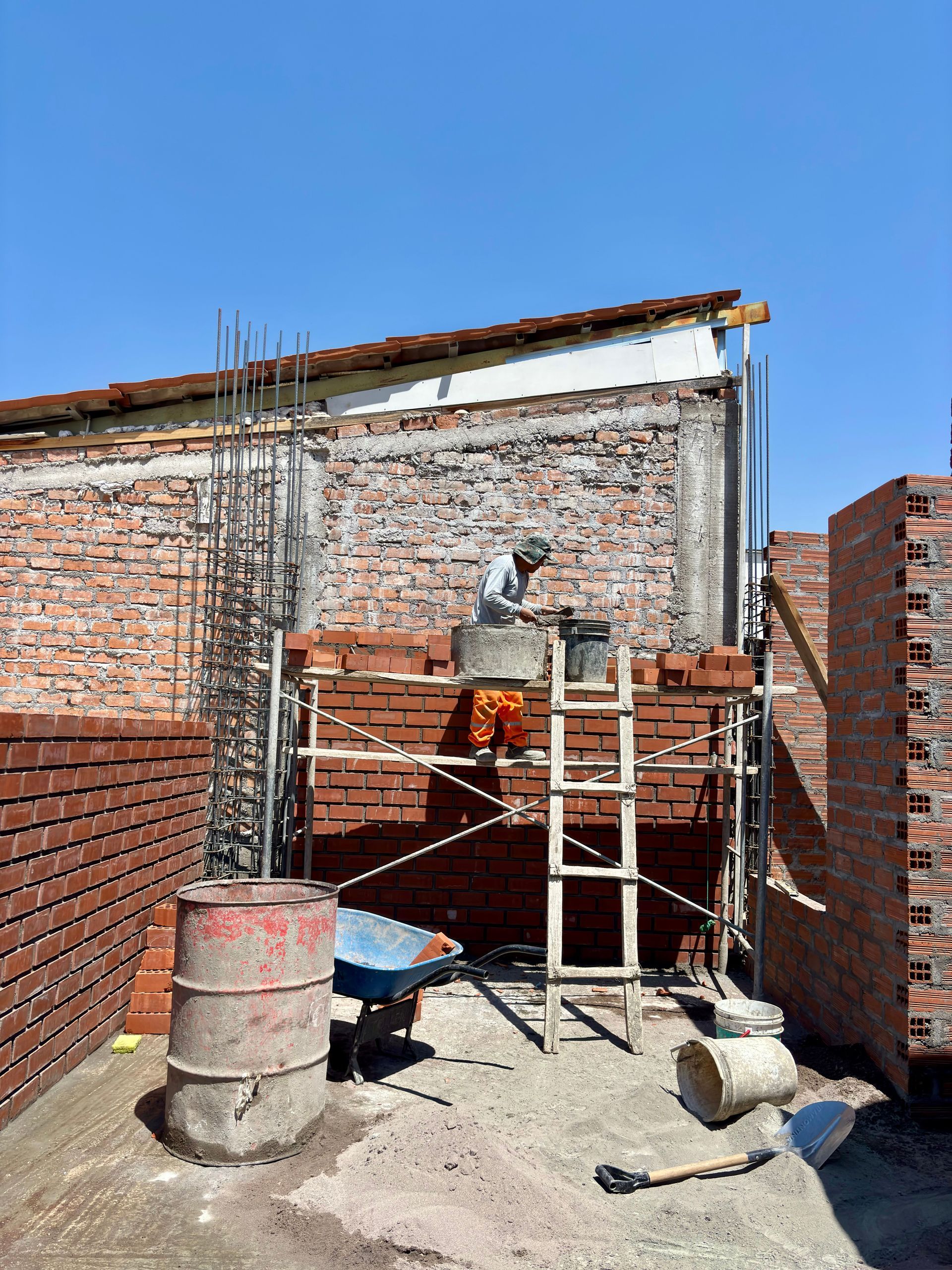 Muro de ladrillo en construcción, con andamios, un barril de cemento y escombros en una zona exterior soleada