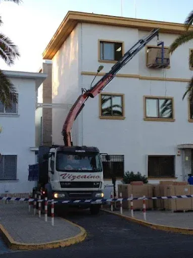 Camión grúa izando materiales frente a un edificio de apartamentos blanco, con cajas de cartón apiladas en el exterior