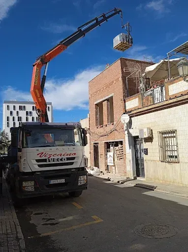 Camión grúa levantando un palé junto a una estrecha calle de ladrillo bajo un cielo azul