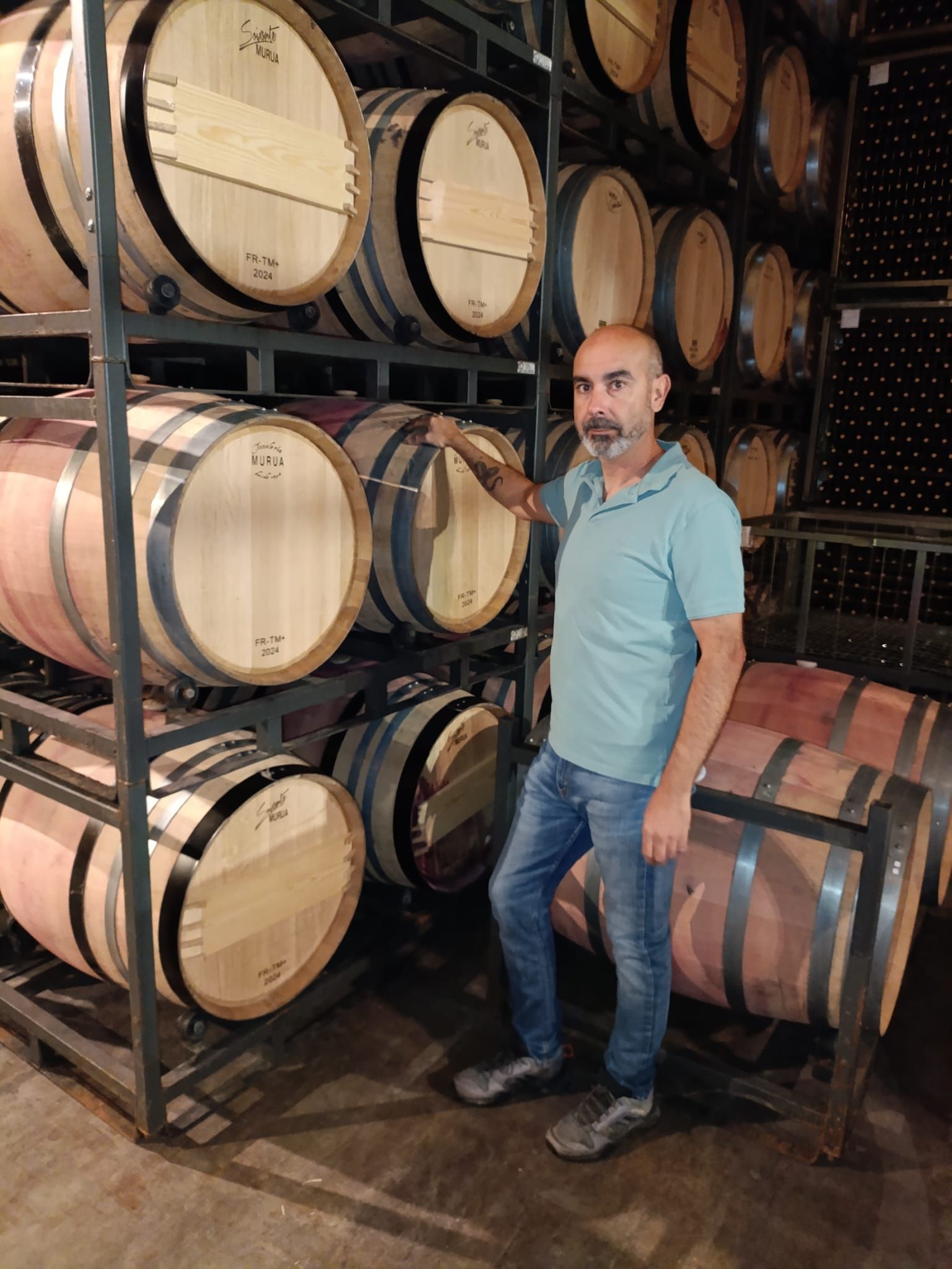 Hombre de pie en una bodega, junto a barriles de madera apilados. Lleva camisa azul y vaqueros.