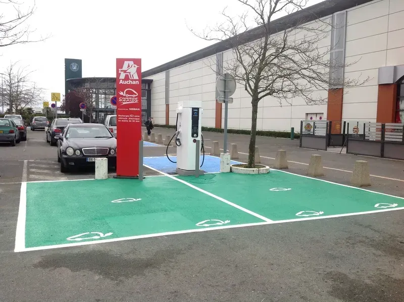Borne de recharge pour véhicules électriques avec un panneau rouge, places de stationnement peintes en vert et une voiture à proximité sur le parking d'un magasin.