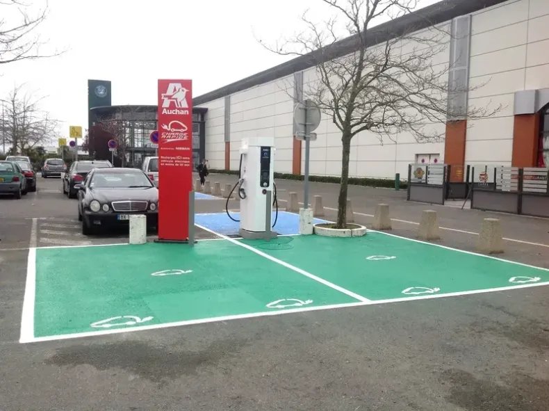Borne de recharge pour véhicules électriques avec un panneau rouge, places de stationnement peintes en vert et une voiture à proximité sur le parking d'un magasin.