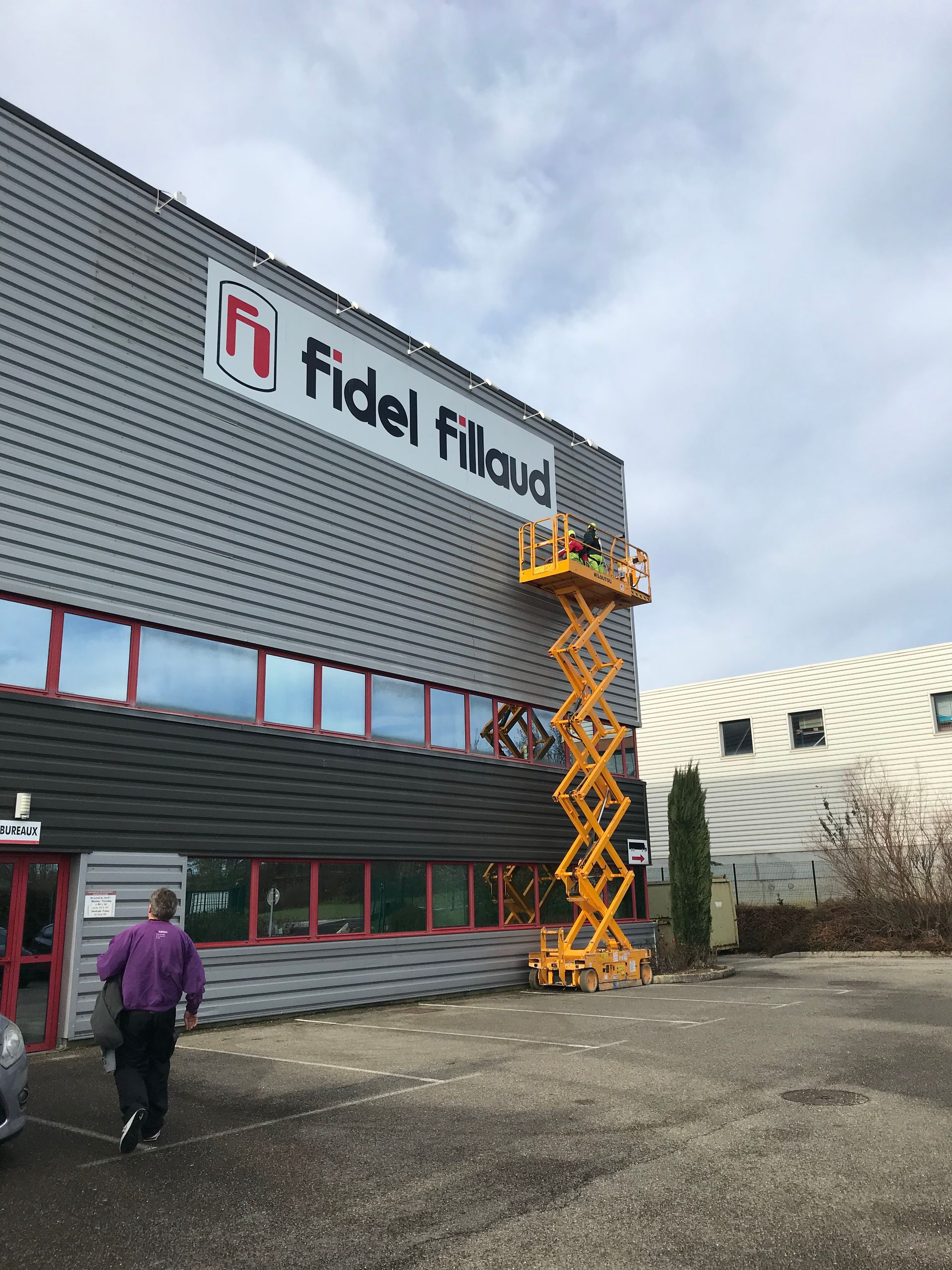 Un ouvrier, à bord d'une nacelle élévatrice jaune, installe une enseigne fidel fillaud, sur la façade d'un bâtiment industriel gris.