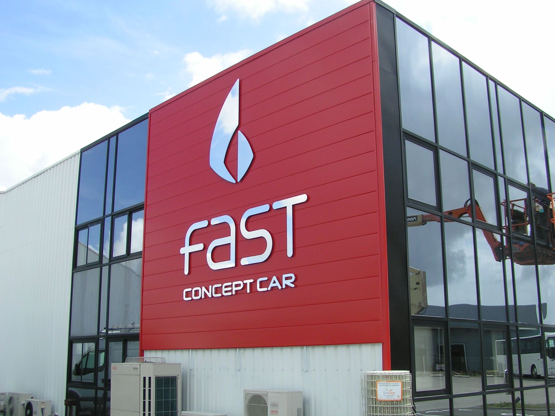 Une façade rouge ornée du logo blanc FAST CONCEPT CAR et mur vitré avec une personne dans une nacelle à coté.