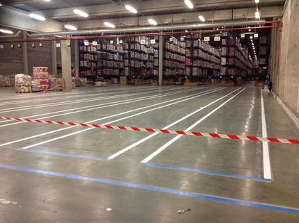 Un entrepôt industriel avec des marquages ​​blancs au sol, des rayonnages à palettes à l'arrière et une barrière de sécurité en ruban adhésif rouge et blanc.