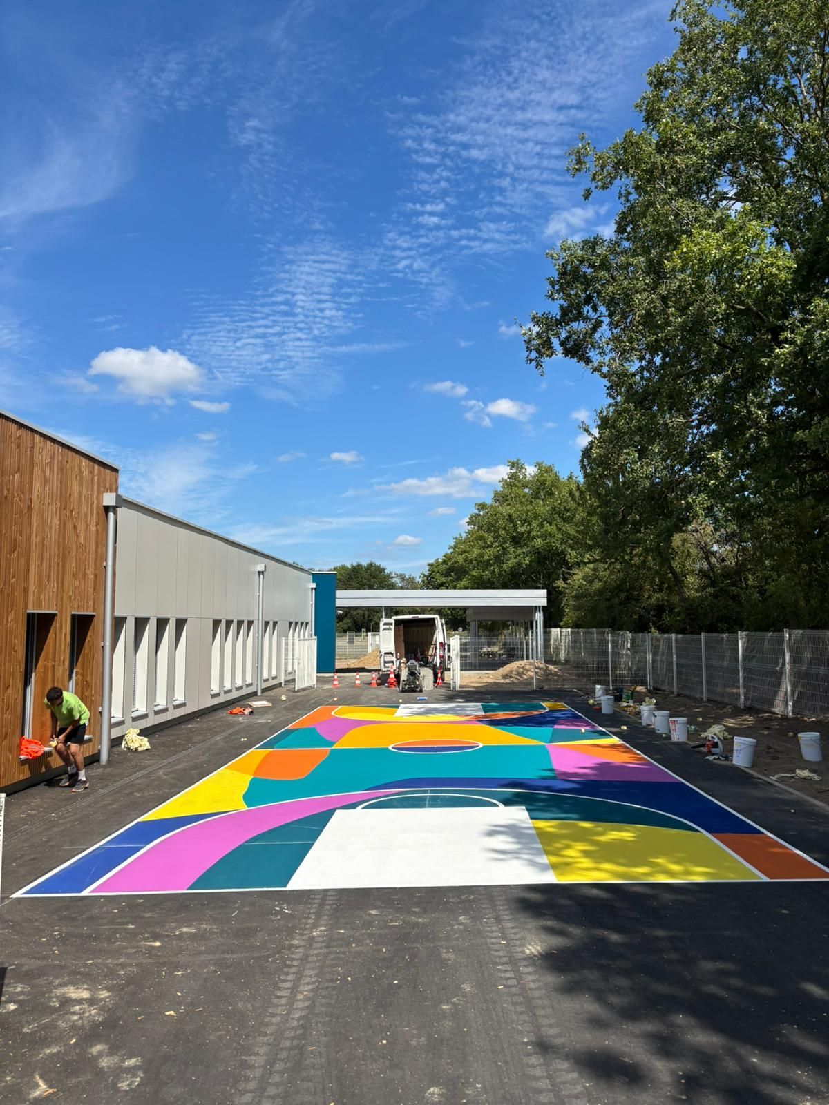 Un ouvrier peint un terrain de basket-ball coloré et des formes géométriques, à côté d'un bâtiment moderne, sous un ciel bleu.