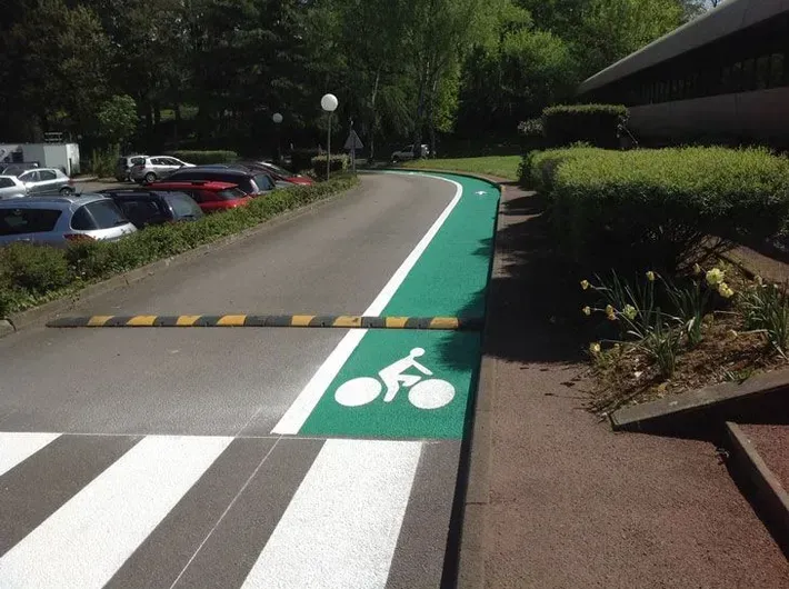 Une piste cyclable peinte en vert et marquée d'un symbole cycliste blanc, longeant un passage piéton et un ralentisseur dans une zone de stationnement.