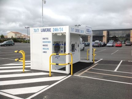 Une laverie automatique extérieure en libre-service avec machines à laver, située sur un parking sous un ciel nuageux.