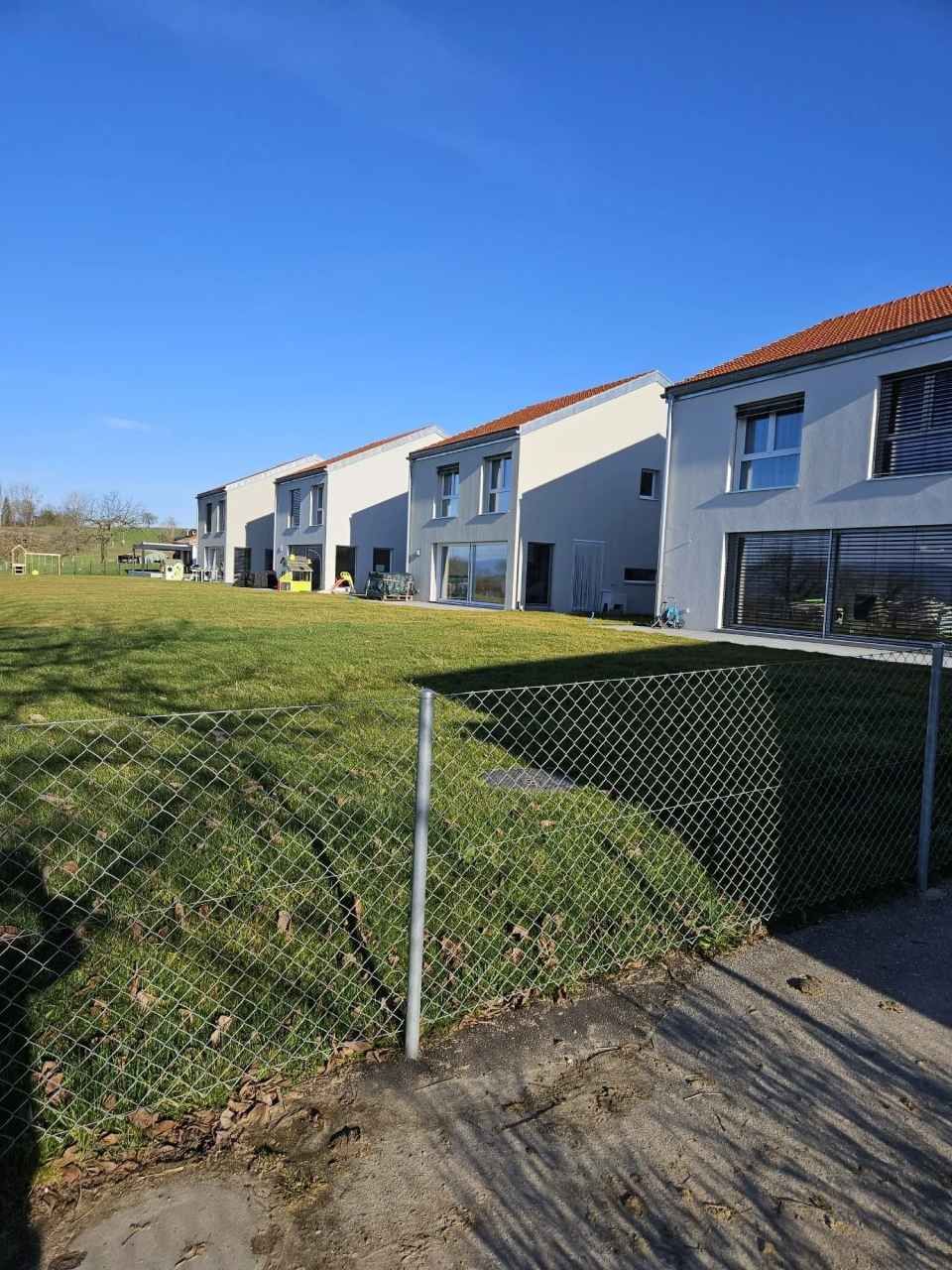 Rangée de maisons modernes aux toits rouges et aux murs gris, derrière une clôture en grillage.