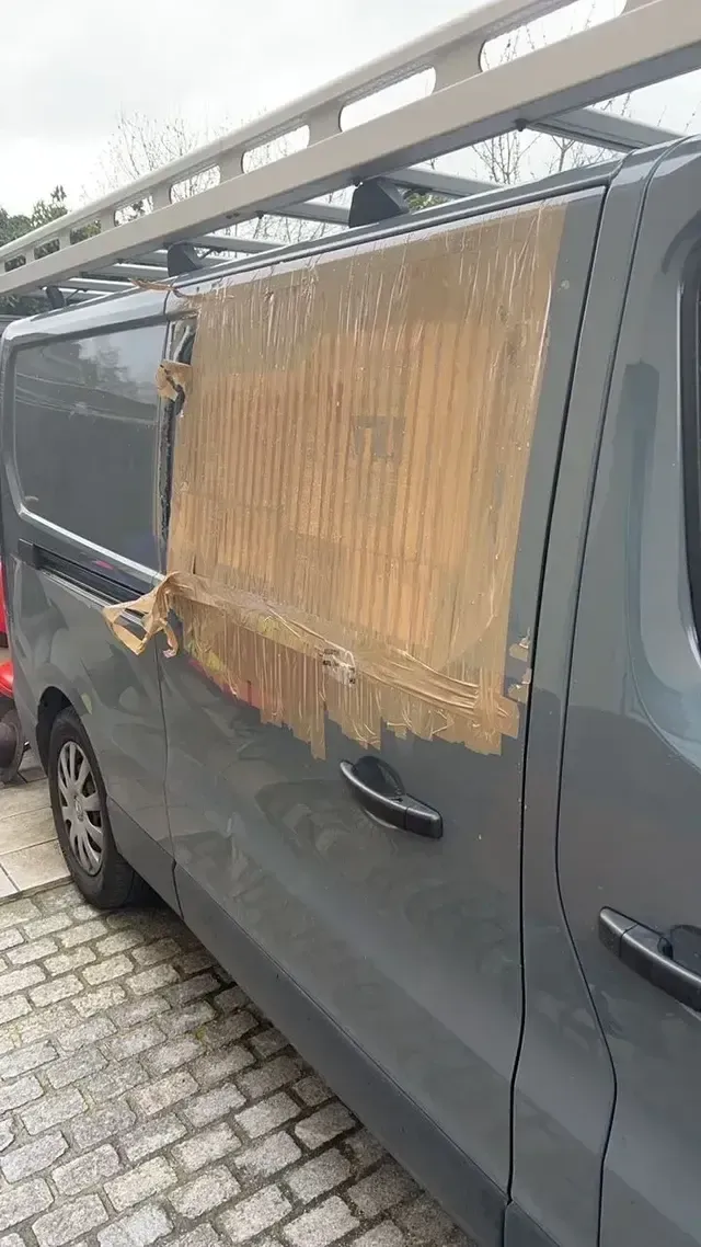 Fourgon gris avec un tapis de bambou scotché couvrant la vitre latérale arrière, garé dans une rue pavée sous un auvent.