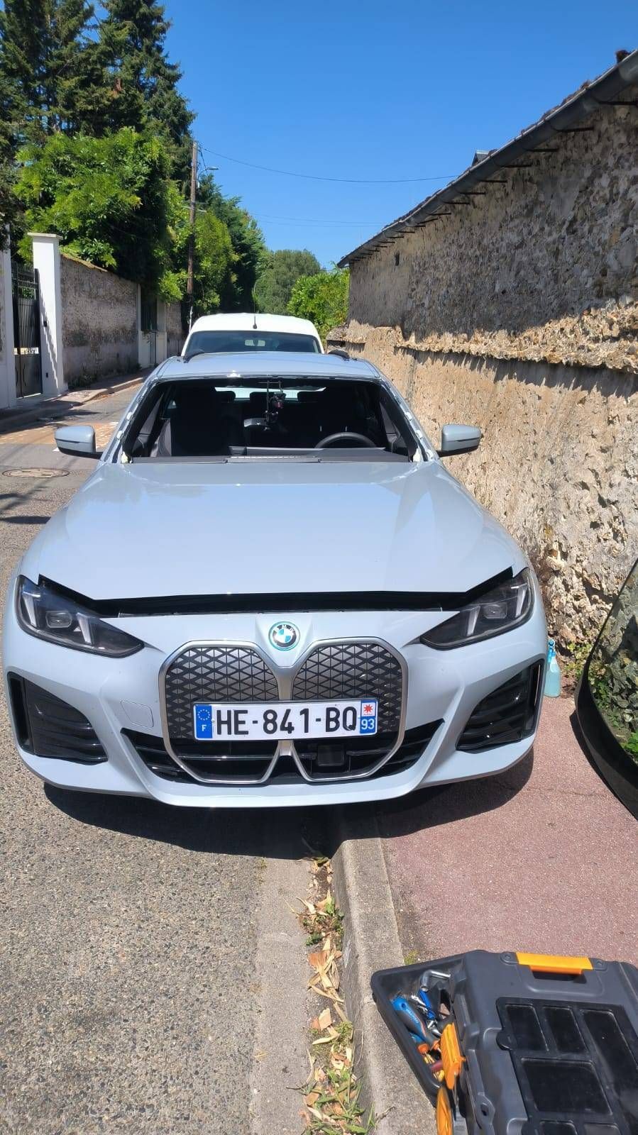 Une voiture blanche est garée dans une rue étroite, près d’un mur en pierre, sous un ciel bleu sans nuages.