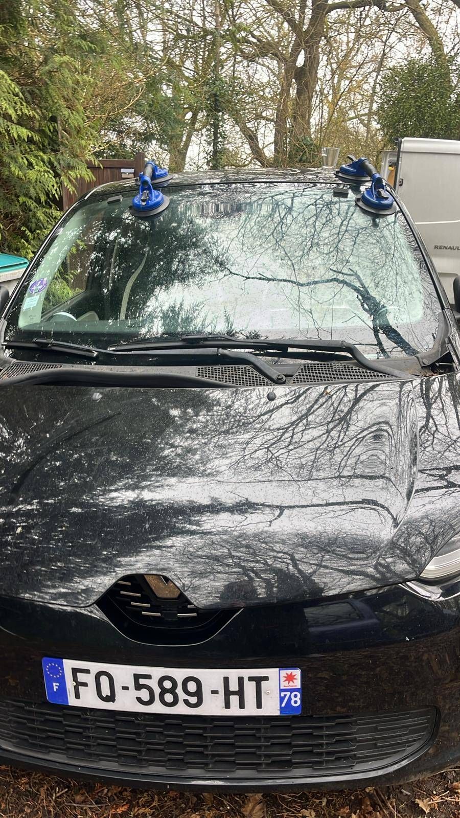 Voiture noire endommagée, avec le pare-brise brisé et deux supports à ventouse bleus sur le toit, garée à l’extérieur