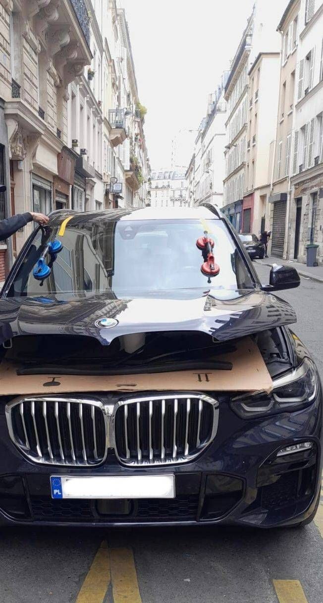 BMW noire avec le capot endommagé, garée dans une rue étroite bordée d’immeubles des deux côtés