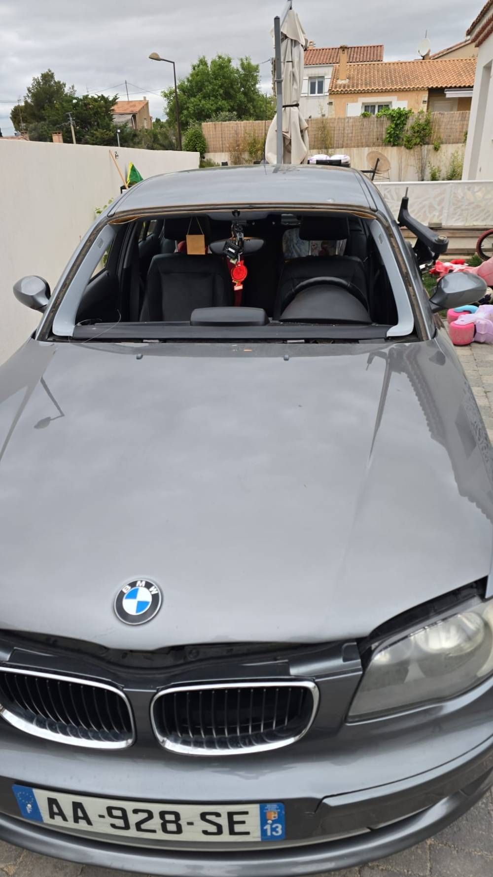 BMW grise avec le pare-brise brisé, stationnée à l’extérieur, avec des objets visibles à l’intérieur.