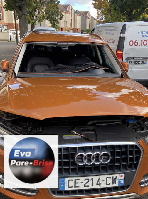 SUV Audi orange avec le pare-brise brisé, garé dans une rue près d’un panneau et d’un arbre