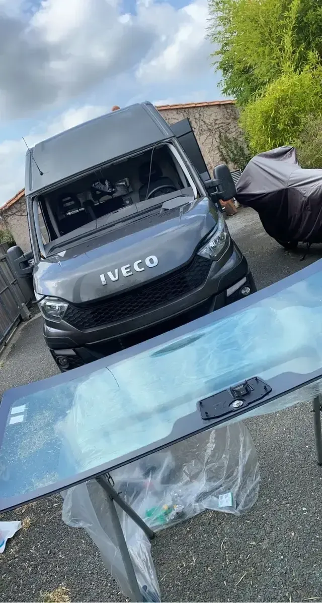 Fourgon IVECO bleu endommagé, avec le pare-brise brisé, garé à côté d’une voiture bâchée dans une allée