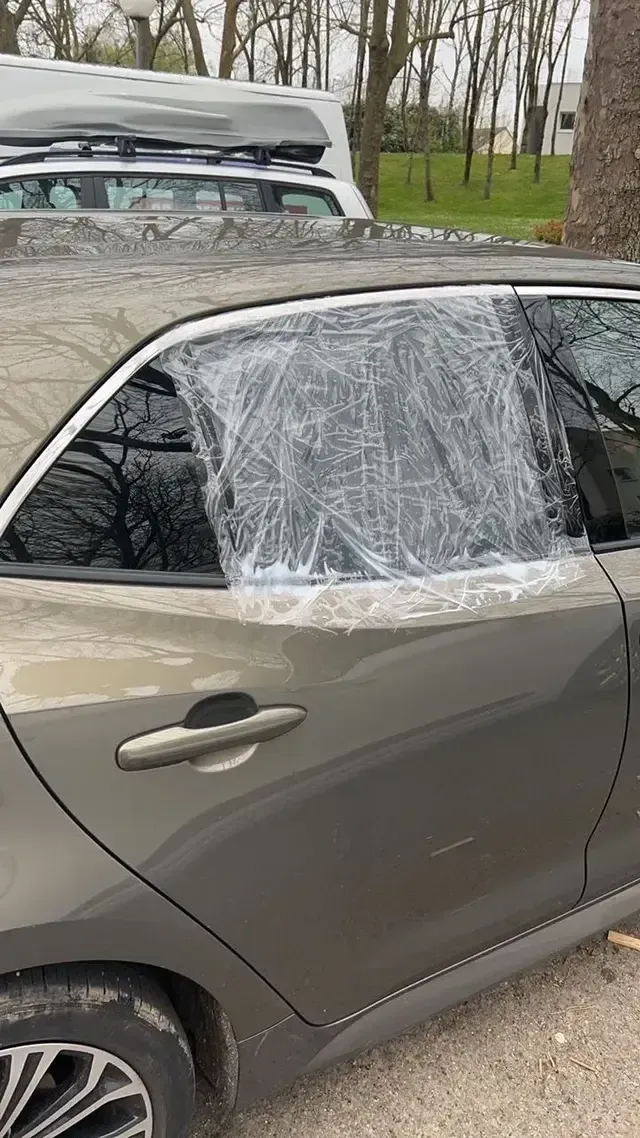 Voiture avec la vitre latérale arrière brisée et recouverte de film plastique transparent, dans une zone boisée