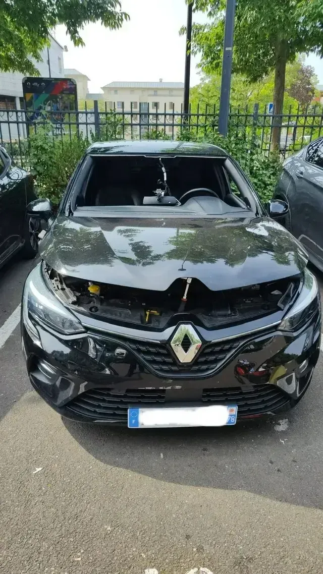 Voiture Renault noire avec le pare-brise et le toit endommagés, garée entre deux voitures à l’extérieur