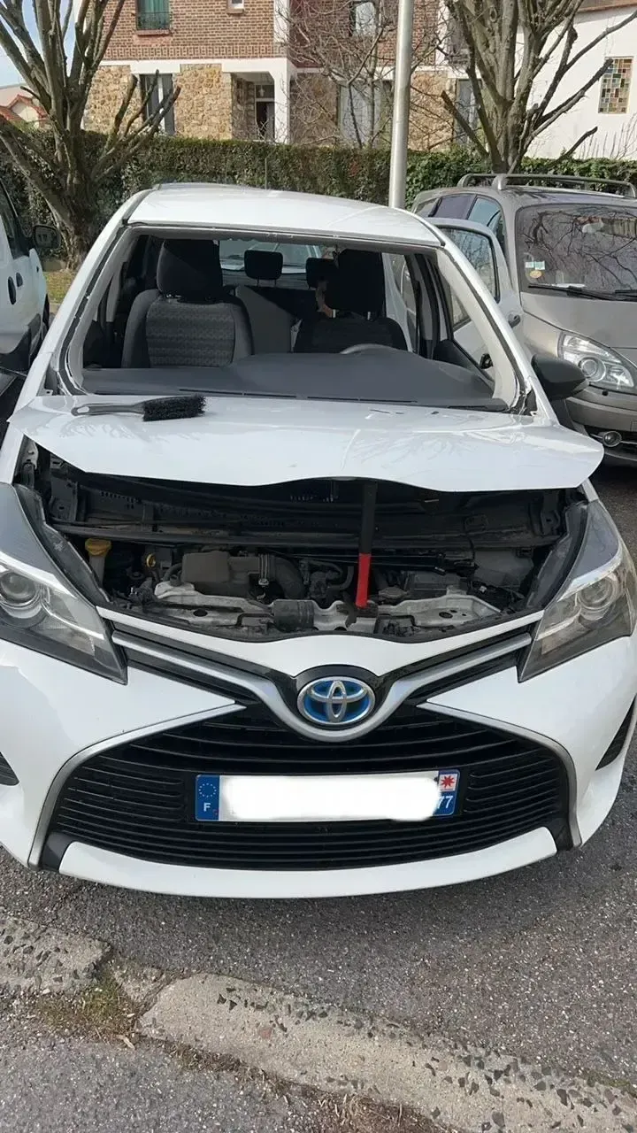 Vue de face d’une Toyota blanche avec le capot endommagé et le pare-brise brisé, garée dans une rue.
