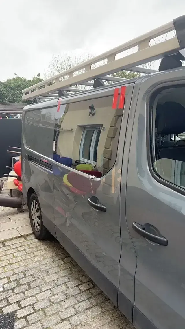 Fourgon gris avec une échelle de toit, garé dans une rue pavée, avec un sac à outils rouge visible à l’intérieur.