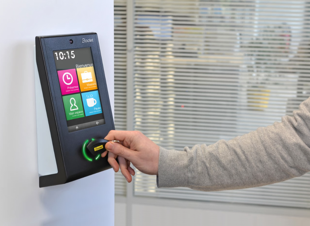 Persona usando una tarjeta de acceso en un reloj de control de entrada en una oficina. La pantalla muestra iconos para marcar entrada y salida.