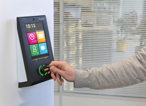 Persona usando una tarjeta de acceso en un reloj de control de entrada en una oficina. La pantalla muestra iconos para marcar entrada y salida.
