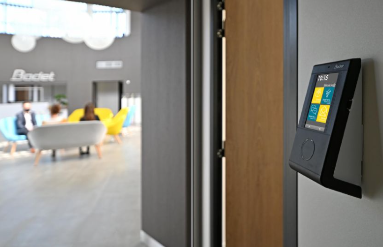 Dispositivo de control de acceso negro en la pared con pantalla colorida, puerta de entrada al vestíbulo de la oficina con gente.