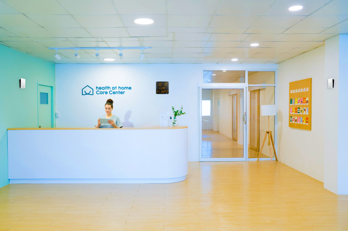 Área de recepción: mujer en un escritorio curvo blanco, logotipo en la pared, piso de madera clara, puerta de vidrio al pasillo.