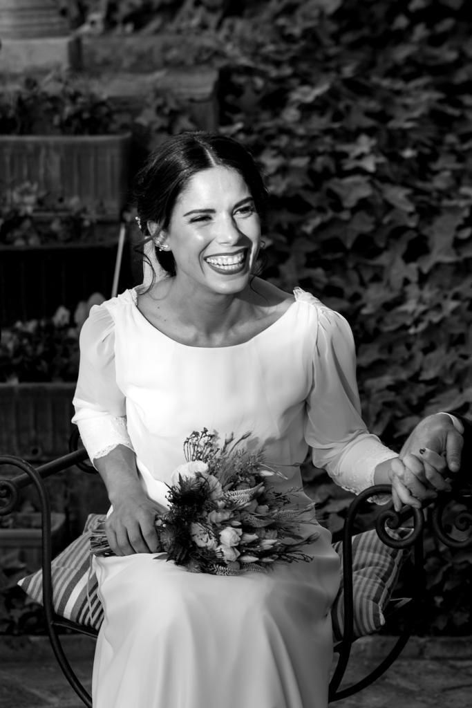 Novia sentada en el jardín, sonriendo, sosteniendo flores, vistiendo un vestido blanco.