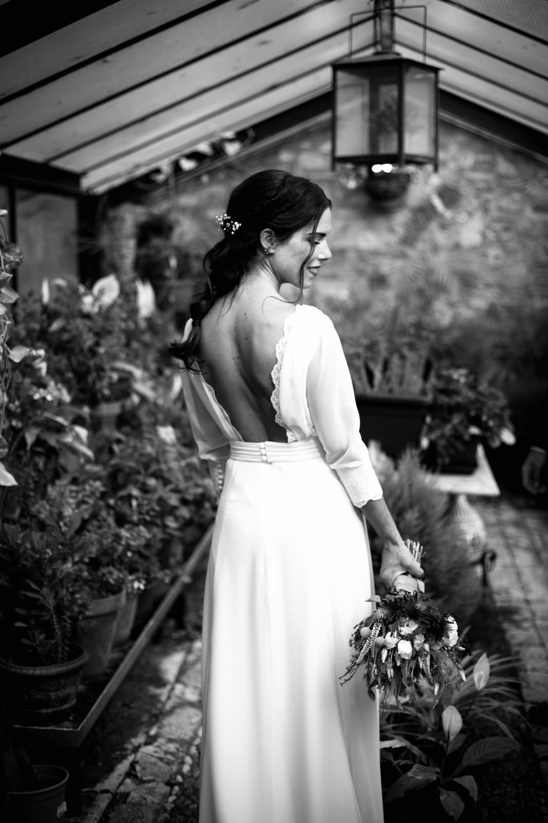 Novia con un vestido blanco, sosteniendo flores, mirando por encima del hombro en un invernadero lleno de plantas.