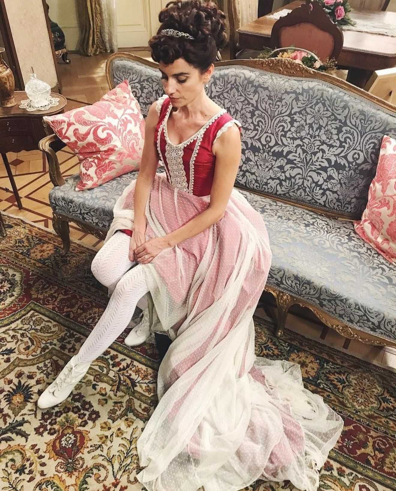 Una mujer con un vestido rojo y blanco está sentada en un sofá azul adornado. Lleva un recogido alto y medias blancas.