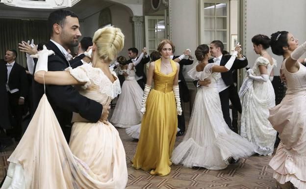 Escena de salón: Bailarines con atuendo formal. Una mujer con vestido amarillo observa a las parejas bailar el vals. Salón elegante.