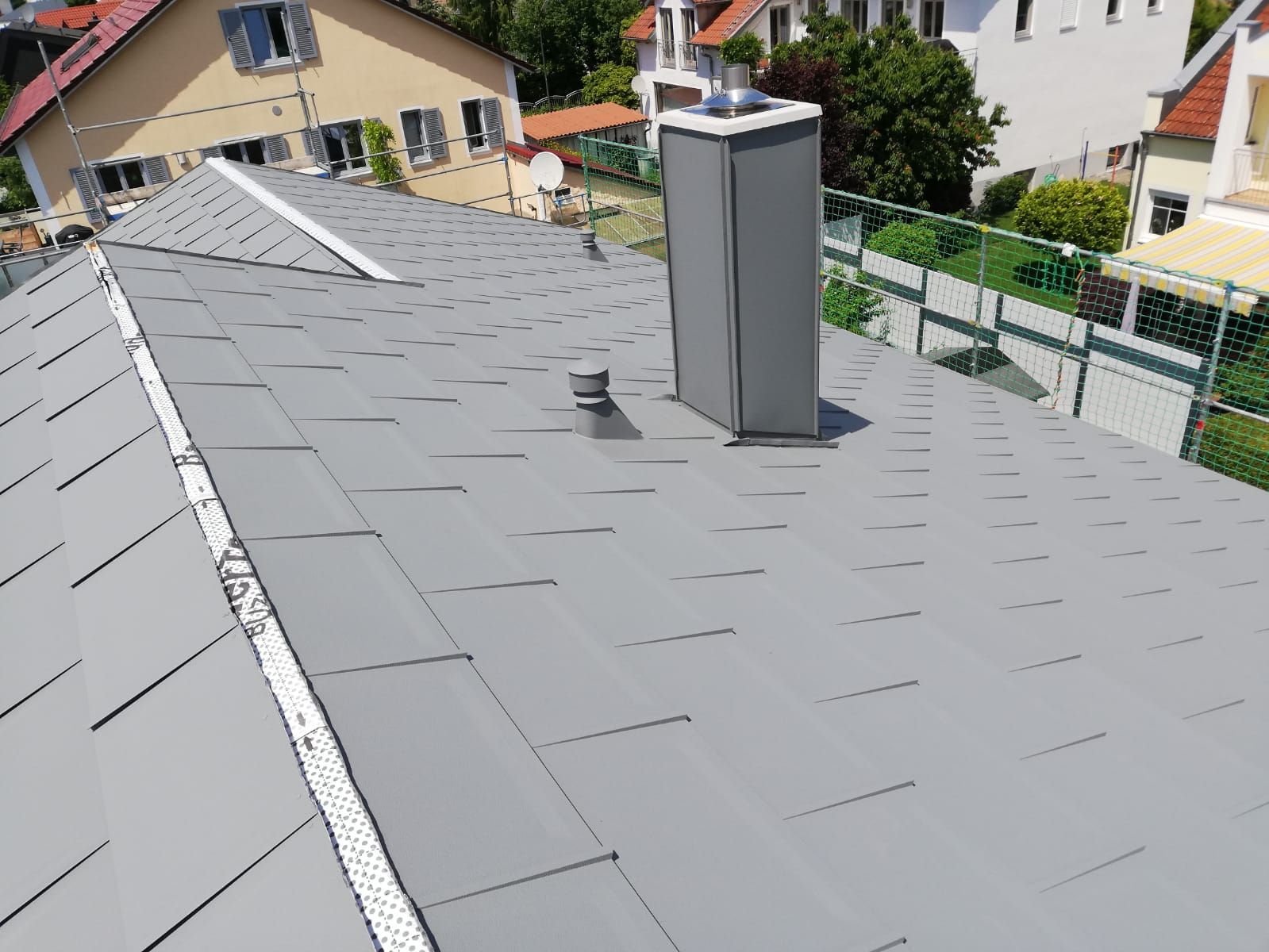 Weitwinkelaufnahme eines grau gedeckten Satteldachs mit einem Schornstein vor einer Wohnsiedlung.