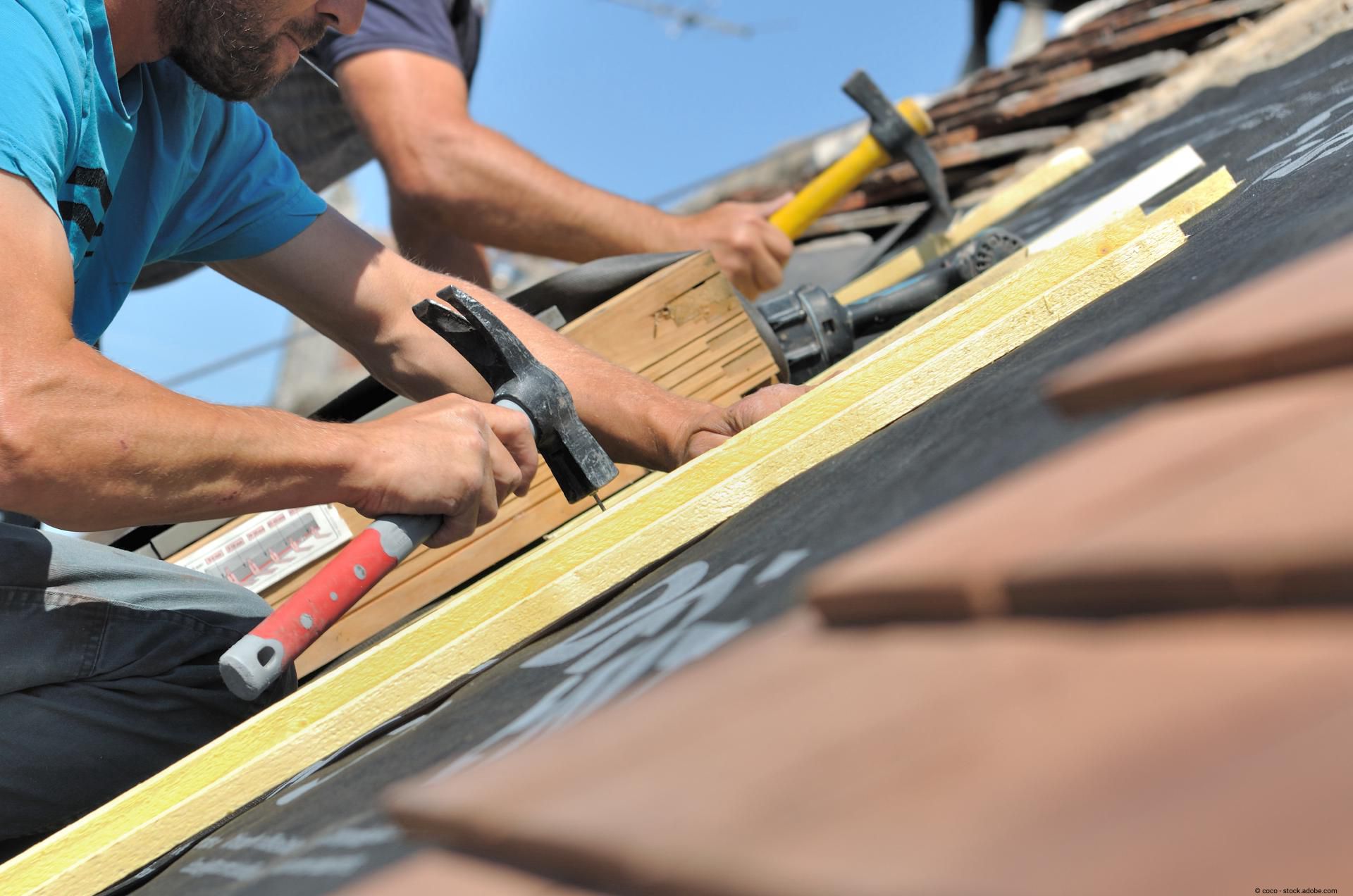 Nahaufnahme eines Handwerkers, der mit einem Hammer Dachlatten auf einer schrägen Dachkonstruktion befestigt.