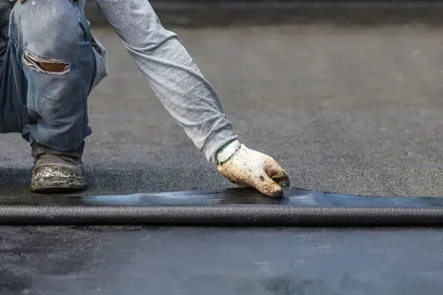 Un trabajador con pantalones vaqueros azules extiende asfalto negro en una carretera con una herramienta manual.