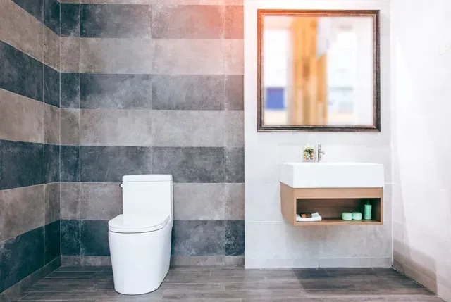 Baño moderno con inodoro, lavabo de madera, espejo y pared de azulejos grises.