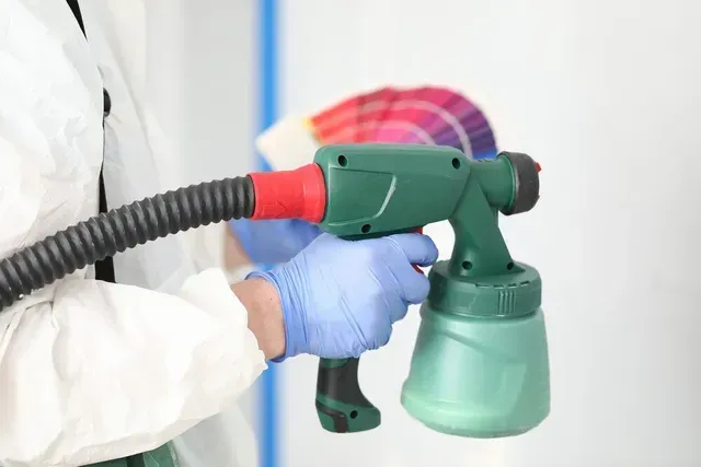 Un trabajador con traje protector rocía una pared con una pistola de pintura verde y guantes azules.