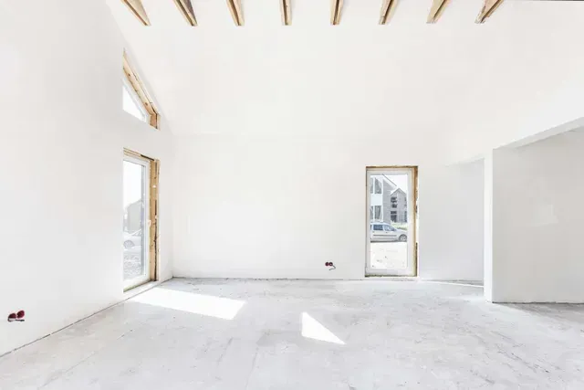 Habitación blanca luminosa y vacía con ventanas altas y una puerta, suelo iluminado por el sol, interior sin terminar.