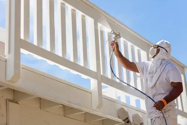 Un trabajador pinta con aerosol una barandilla blanca en un balcón bajo un cielo azul despejado.