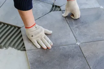 Manos enguantadas instalando grandes baldosas grises de patio sobre una superficie de piedra exterior.