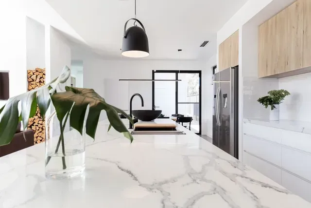 Cocina moderna y luminosa con isla de mármol, lámpara colgante y sala de estar abierta.