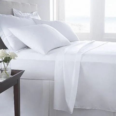 Una cama con sábanas y almohadas blancas y un jarrón de flores en una mesita de noche.