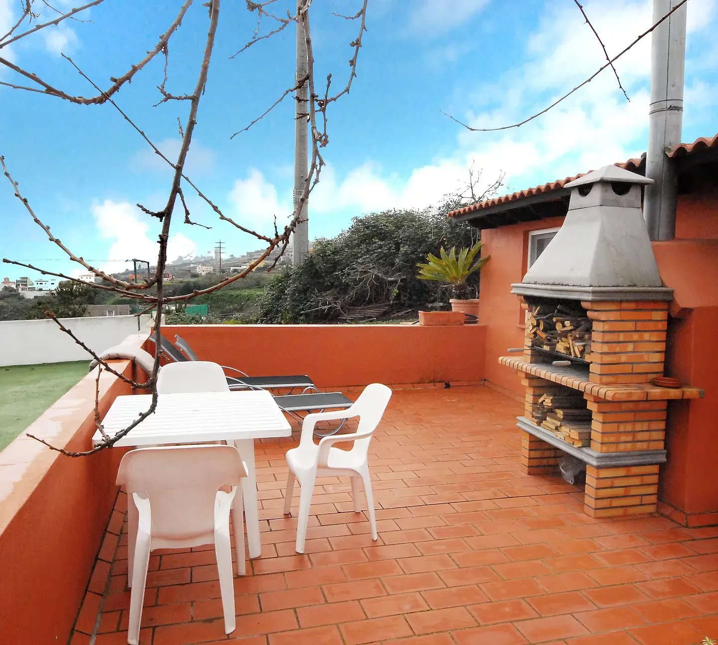 Un soleado patio exterior de terracota con una mesa blanca con sillas, tumbonas y una chimenea de piedra con barbacoa.